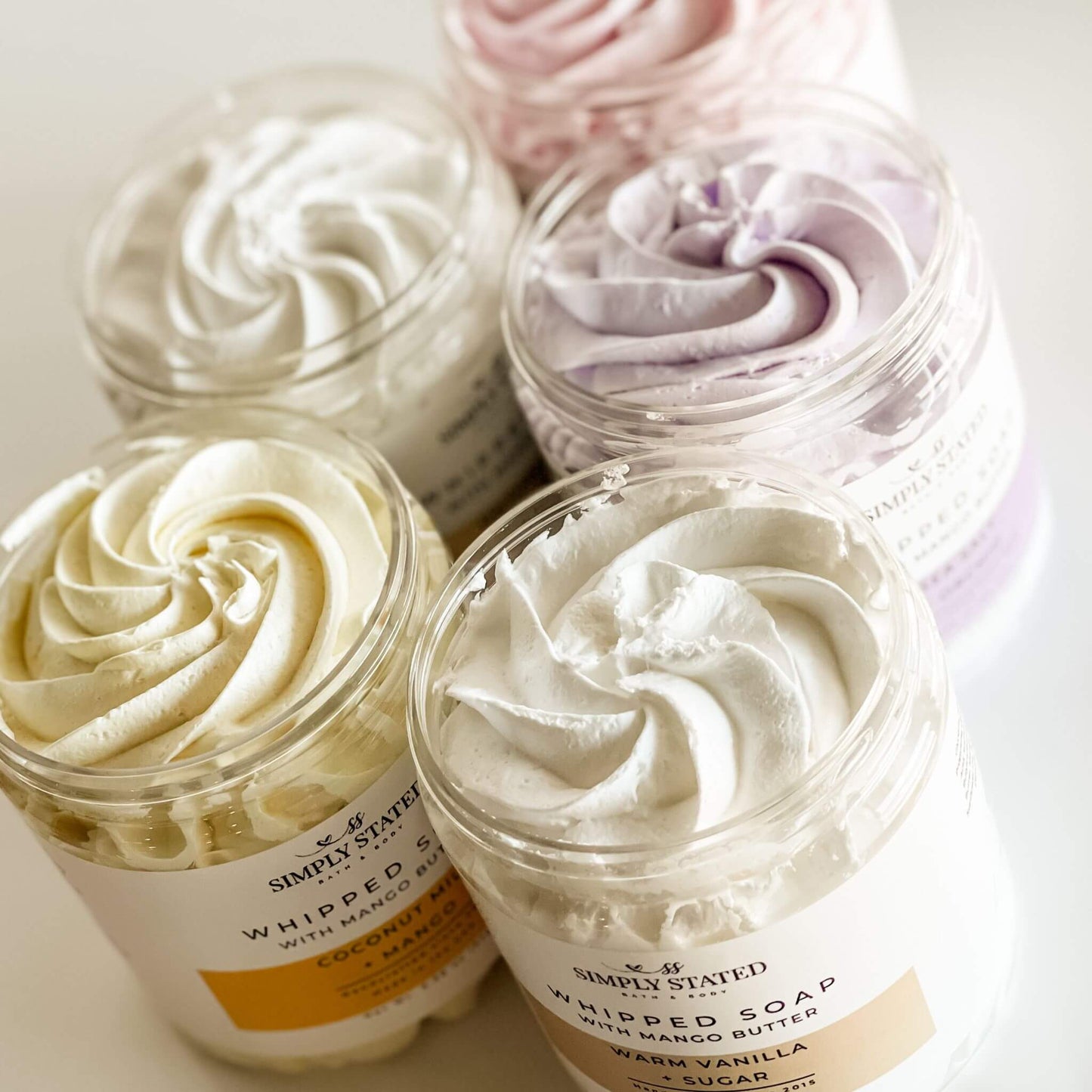 Open jars of Signature Line Whipped Soaps showcasing creamy whipped textures in vibrant colors, including Coconut Milk + Mango, Sweet Berry + Vanilla, Warm Vanilla + Sugar, Oatmeal Milk + Honey, and Sea Salt + Orchid.