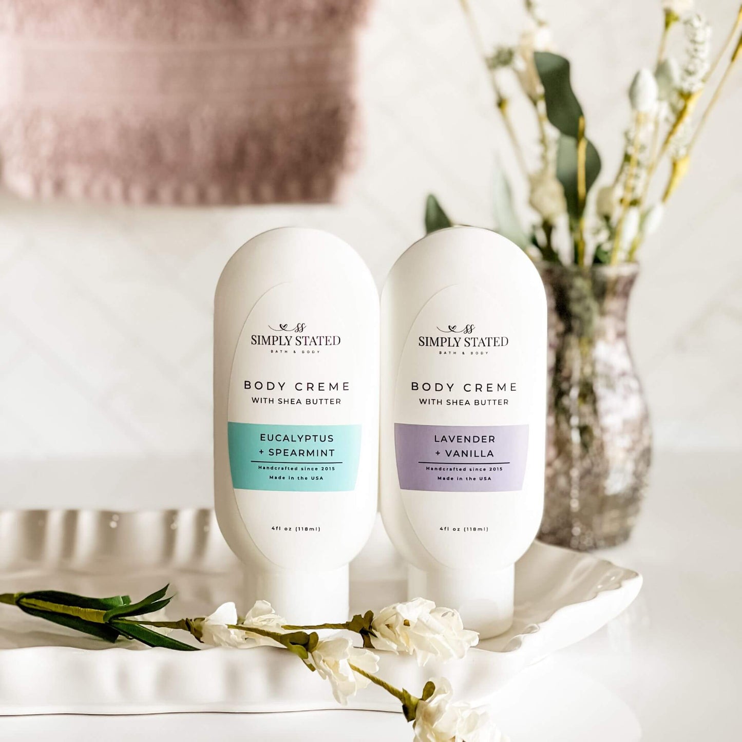 Two bottles of Simply Stated Bath & Body Aromatherapy Body Creme in Eucalyptus + Spearmint and Lavender + Vanilla, displayed on a white tray with floral accents. A soft, neutral background enhances the calming aesthetic.