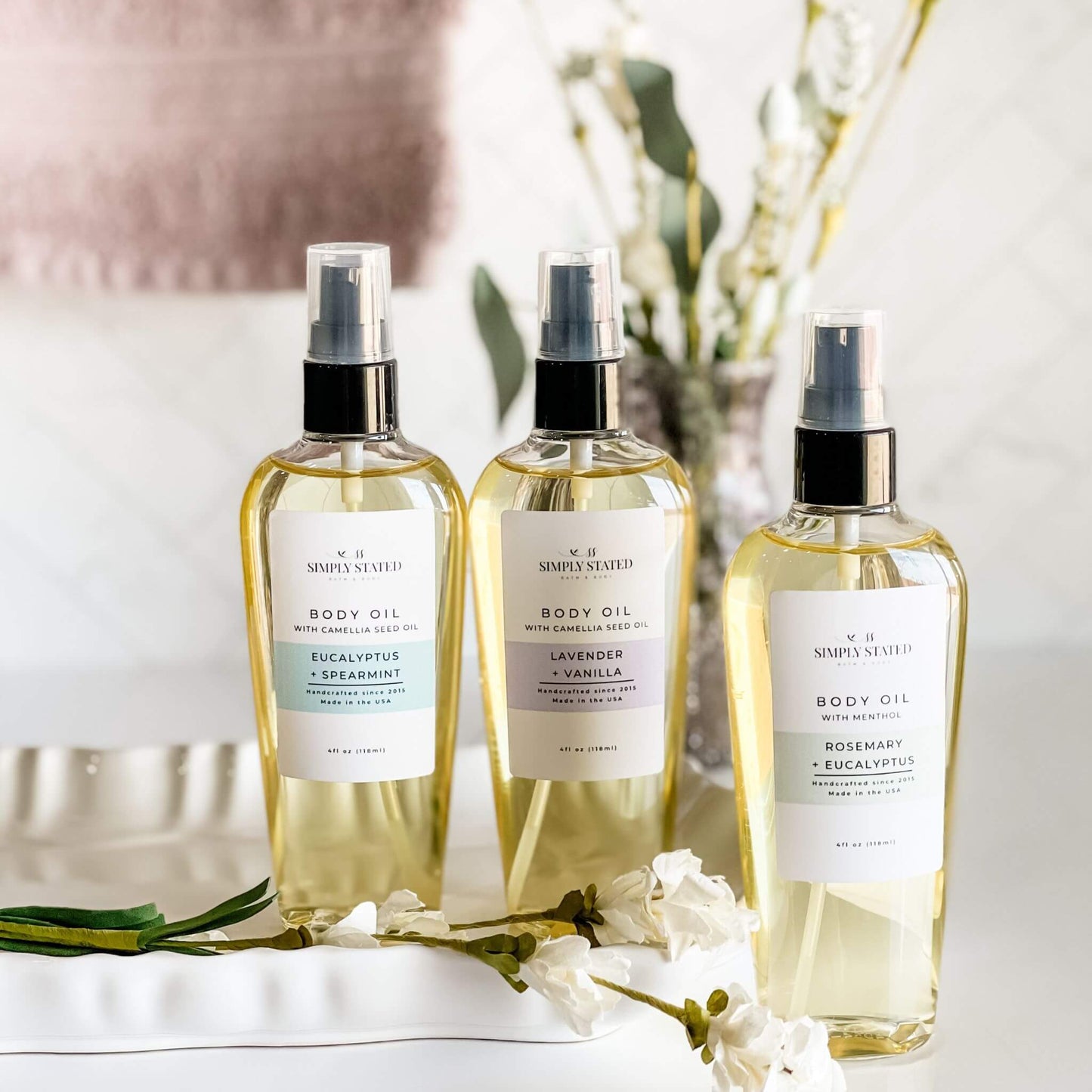 Three bottles of Simply Stated Bath & Body Aromatherapy Body Oil in Eucalyptus + Spearmint, Lavender + Vanilla, and Rosemary + Eucalyptus, arranged on a white tray with elegant floral details and a neutral, spa-inspired backdrop.