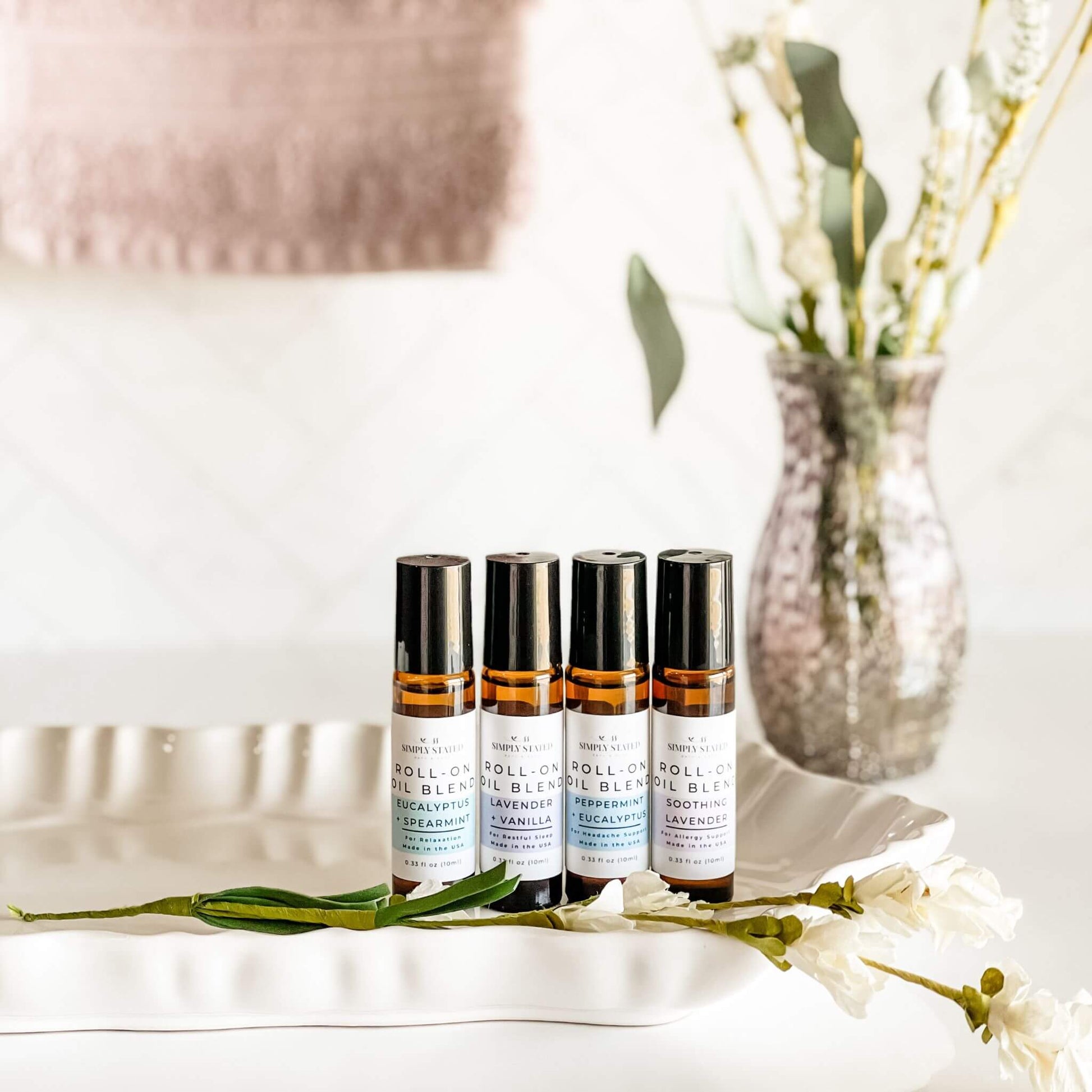 Four Simply Stated Bath & Body Roll-On Oil Blends, featuring Eucalyptus + Spearmint, Lavender + Vanilla, Peppermint + Eucalyptus, and Soothing Lavender. Arranged on a white tray with a calming floral aesthetic.