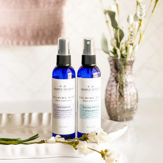 Two bottles of Simply Stated Bath & Body Calming Mist in Eucalyptus + Spearmint and Lavender + Chamomile, elegantly displayed on a white tray with fresh greenery and neutral decor, embodying relaxation and wellness.
