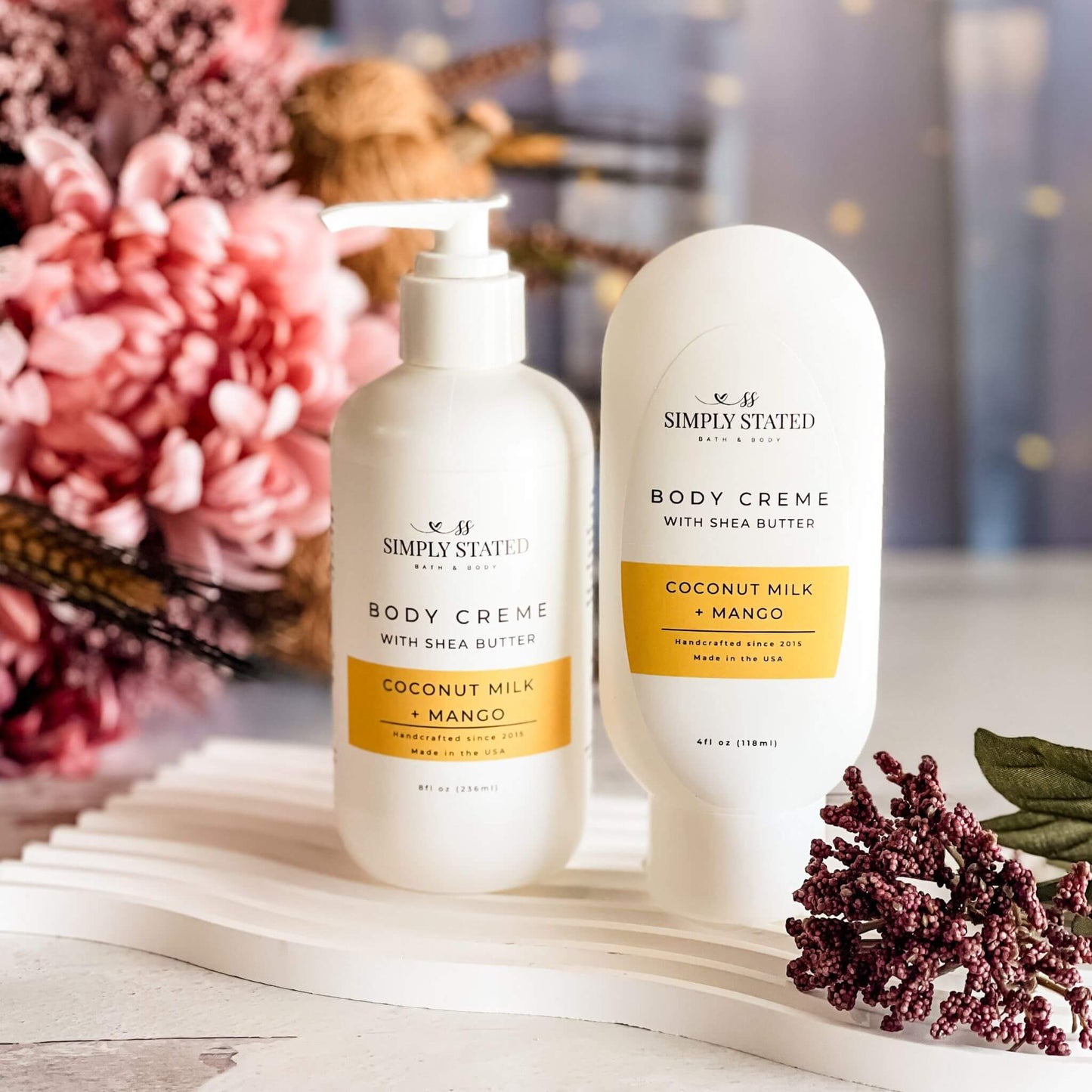 Two bottles of Simply Stated body cream on a decorative surface with flowers in the background