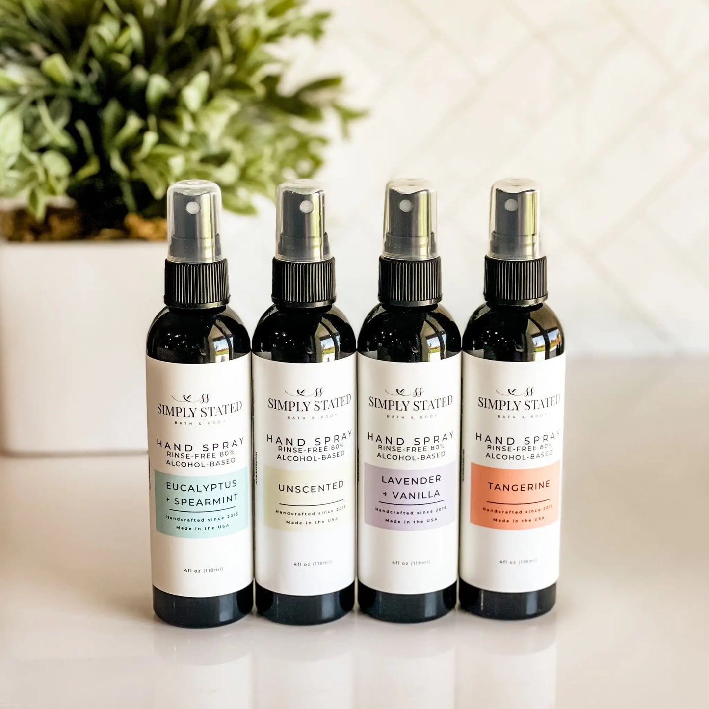 Four bottles of Simply Stated Hand Spray on a light surface with a plant in the background.