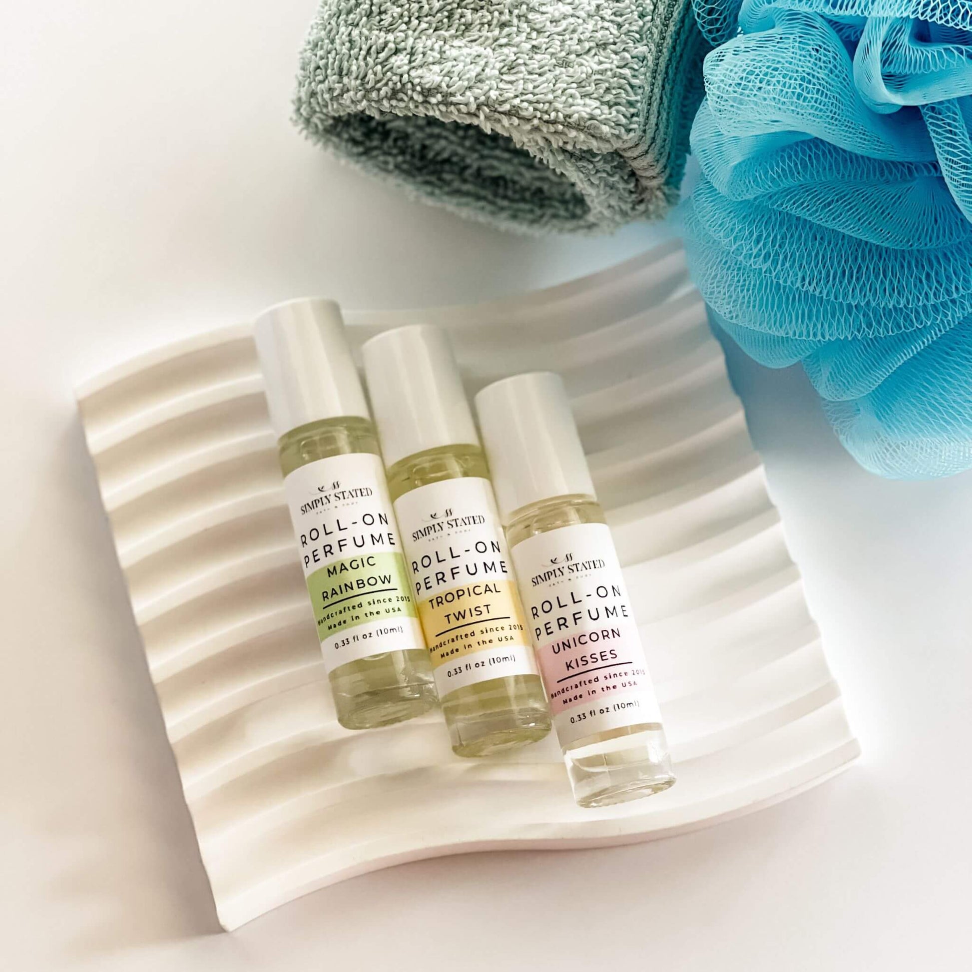 Three roll-on perfume bottles on a textured white plate with a towel and loofah in the background.