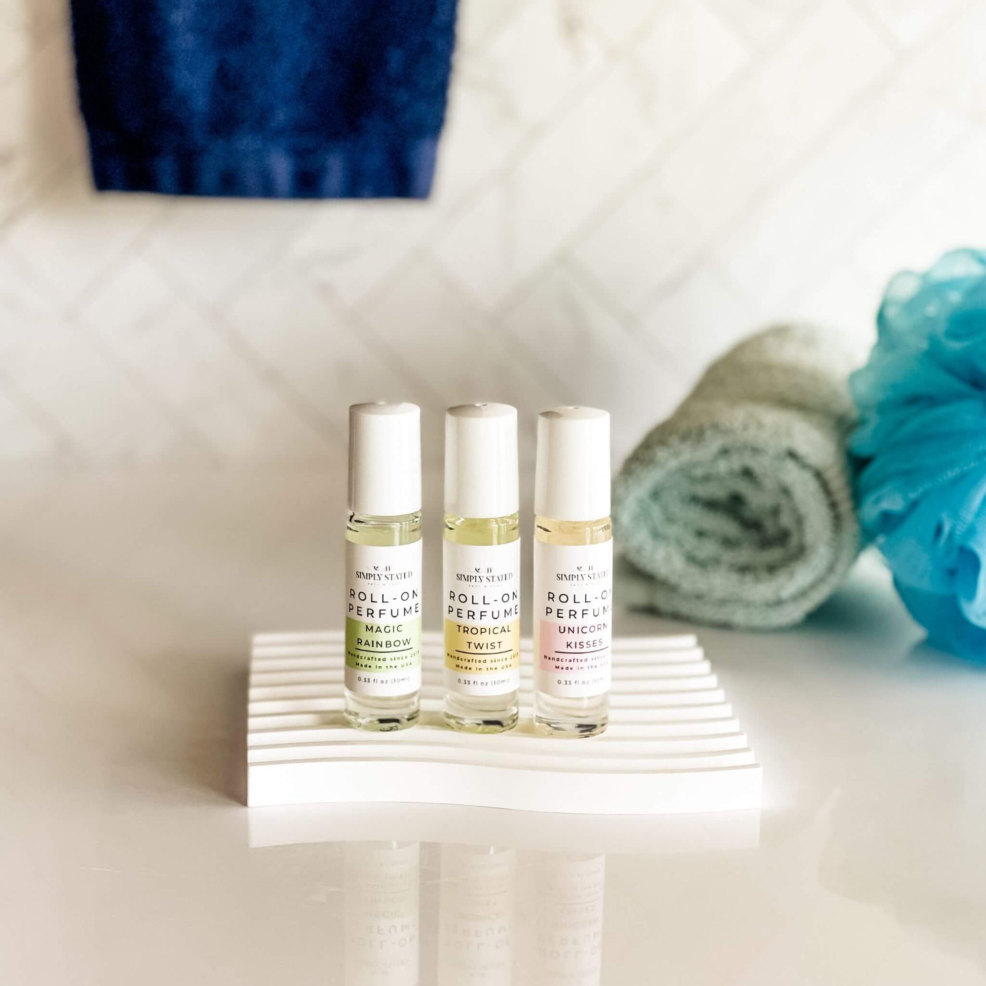 Three small rollerball bottles of roll-on perfume on a white surface with bath towels and a loofah in the background.