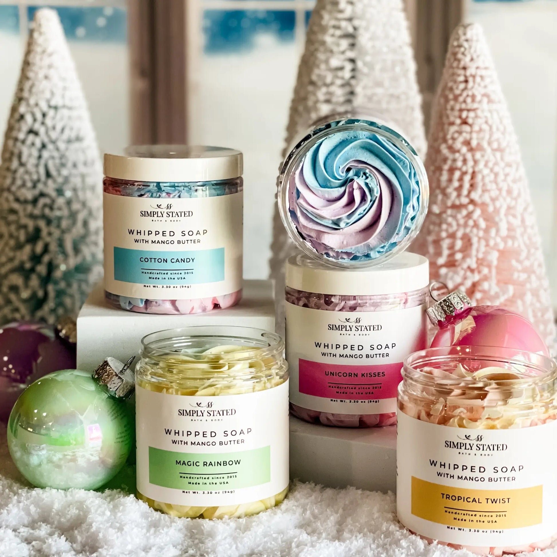 Collection of whipped soap jars with colorful labels and decorative Christmas trees in the background.
