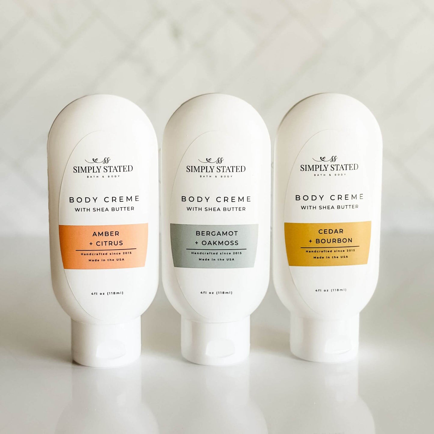 Group of 4 oz men's body creme bottles in three scents: Amber + Citrus, Bergamot + Oakmoss, and Cedar + Bourbon.