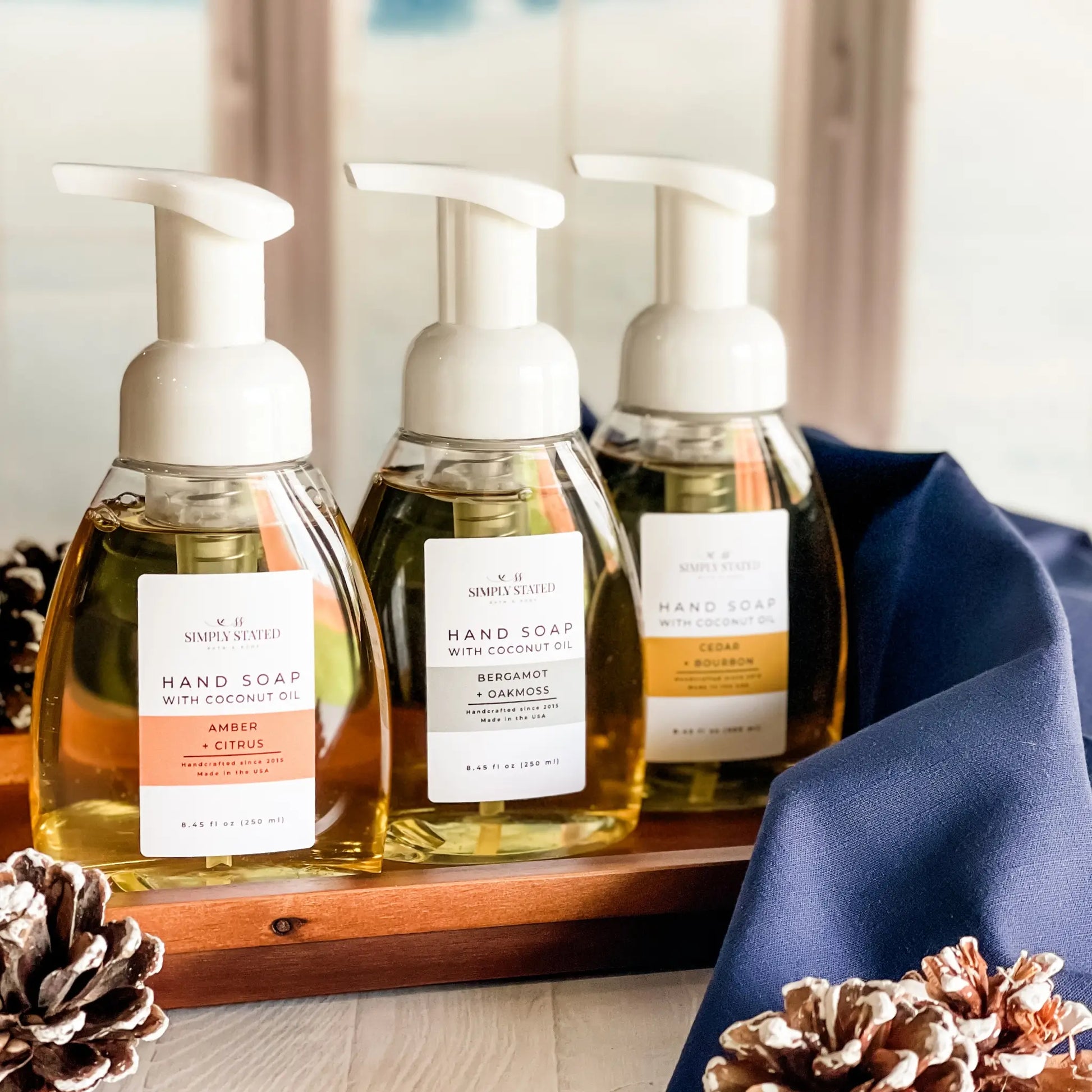 Three bottles of hand soap with pump dispensers on a wooden tray, surrounded by decorative pinecones.