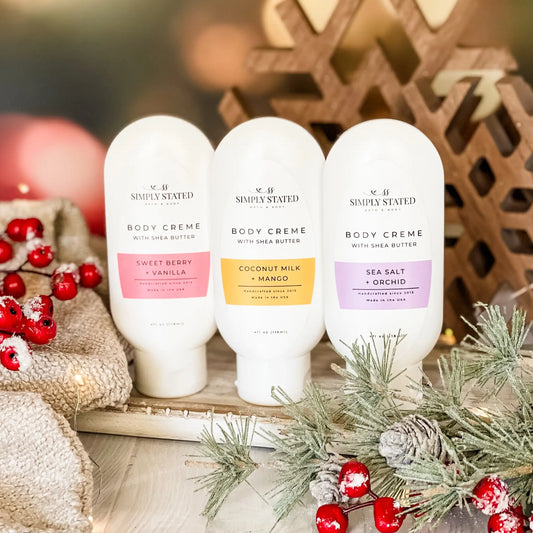 Three bottles of 'Simply Stated' body crème with holiday decorations in the background