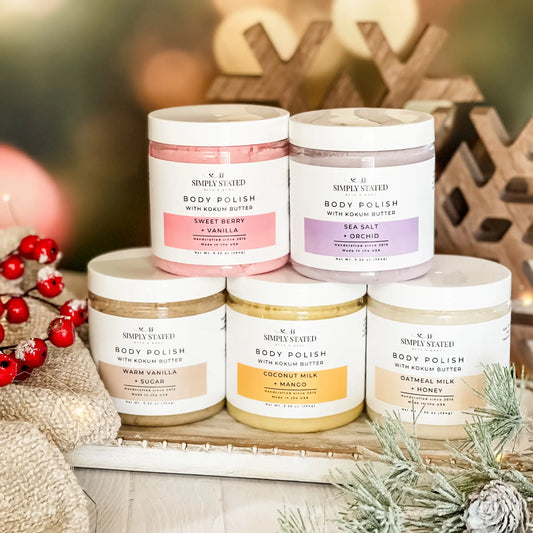 Five jars of body polish with different scents on a decorative surface with Christmas decorations.